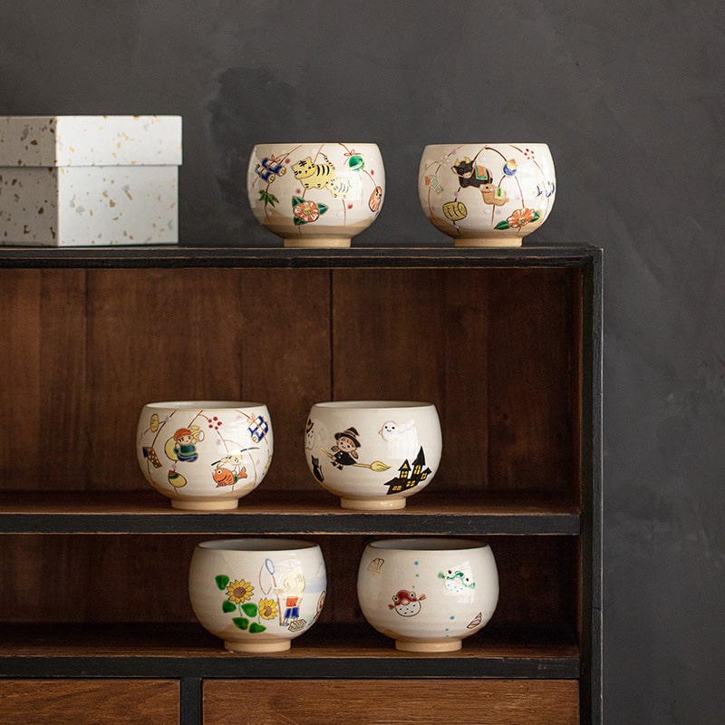 Collection of hand-painted ceramic cups with whimsical designs on a wooden shelf.