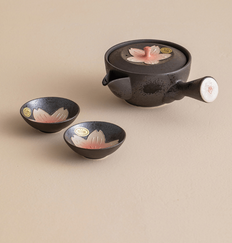 Black kyusu teapot with pink sakura blossom motif and matching teacups on a wooden surface.