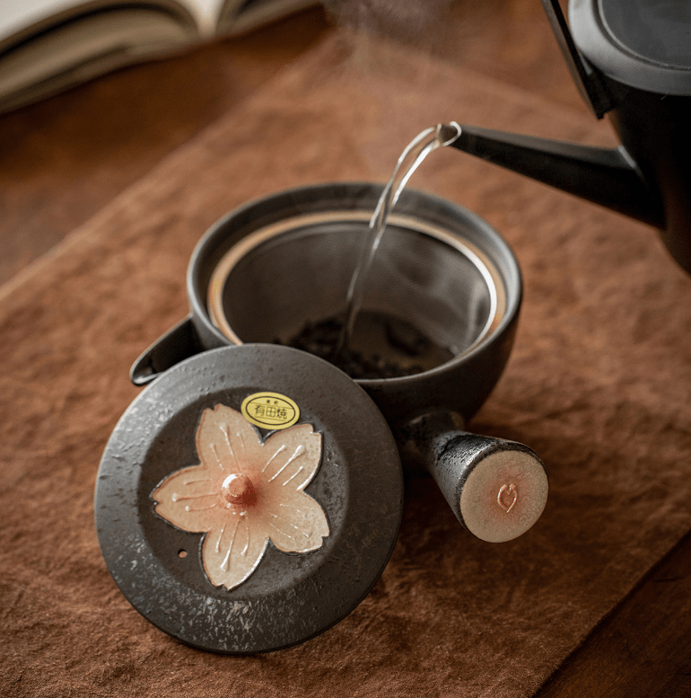 Black kyusu teapot with pink sakura blossom motif and matching teacups on a wooden surface.